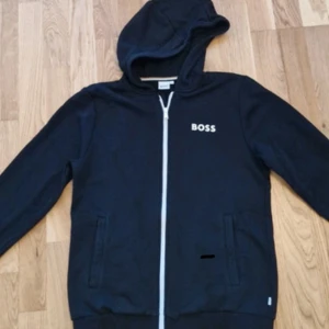 Svart hoodie från BOSS med dragkedja - Svart hoodie från BOSS med vit logga på bröstet och hel dragkedja framtill. Tröjan har huva, två fickor och ribbade muddar vid ärmslut och nederkant. Perfekt för chill dagar och enkel att matcha med jeans eller joggers.