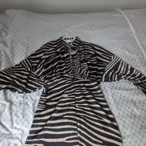 Zebramönstrad kort klänning med krage HM - Snygg kort klänning med zebramönster i svart och beige. Klänningen har en klassisk skjortkrage, knappar framtill och långa ärmar med bred passform. Materialet är glansigt och känns lyxigt mot huden, perfekt för dig som vill sticka ut med ett djuriskt print. Använd en gång.