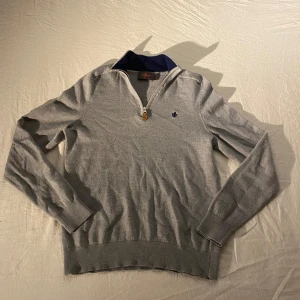Grå merinoulltröja från Morris - Snygg grå långärmad tröja från Morris i 100% merinoull. Tröjan har half zip med läderdetalj och en broderad mörkblå logga på bröstet. Ribbstickade muddar och krage med marinblå insida ger en clean och stilren look.