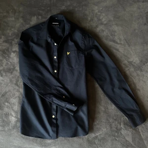Marinblå skjorta från Lyle & Scott - Marinblå skjorta från Lyle & Scott med klassisk krage, vita knappar och bröstficka med gul broderad logga. Skjortan är långärmad och har en stilren, enkel design som passar till många olika looks.