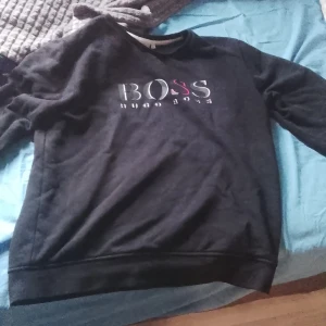 Svart sweatshirt från Hugo Boss - Svart sweatshirt från Hugo Boss med stor BOSS-logga i vitt och rött på bröstet. Tröjan har rund hals och långa ärmar, perfekt för en chill och stilren look. Materialet känns mjukt och skönt mot huden, och passformen är normal. Snygg att matcha med jeans eller joggers.