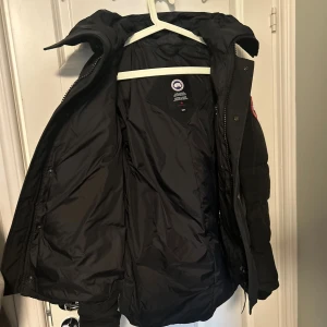 Svart dunjacka från Canada Goose - OBS: Äkta och kvitto finns.                                  Säljer en svart dunjacka från Canada Goose  - Macmillan med huva och klassisk logga på ärmen. Jackan har stora fickor framtill, dragkedja och knäppning. Perfekt för kalla vinterdagar och riktigt snygg streetstil. Säljs då jag växt ur den. KVITTO FINNS, Kan mötas upp i Stockholm eller frakta i Sverige. Väger 82, 176 cm och den passar mig. 
