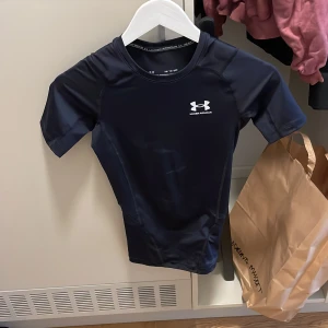 Svart kompressionst-shirt Under Armour - Säljer en svart kompressionst-shirt från Under Armour i storlek XS. T-shirten har korta ärmar, rund halsringning och är tillverkad i ett stretchigt syntetmaterial som andas. Perfekt för träning och sportiga aktiviteter. Diskret logga på bröstet.