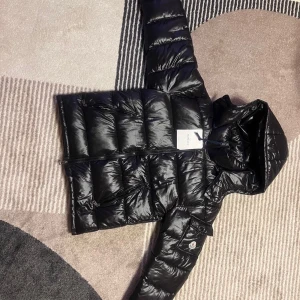 Svart Moncler pufferjacka med huva - Svart pufferjacka från Moncler med glansig finish, huva och dragkedja framtill. Jackan har quiltad design, långa ärmar och Moncler-logga på ärmen. Perfekt för kalla dagar och har en modern, clean look som funkar till streetwear.