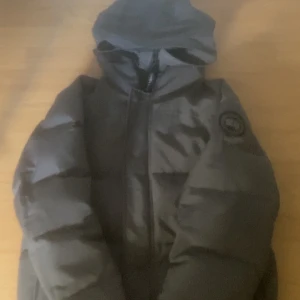 Svart dunjacka från Canada Goose - Säljer en svart dunjacka från Canada Goose i storlek M. Jackan har huva, dragkedja och klassisk patch på ärmen. Fylld med dun och fjädrar för extra värme, perfekt för kalla dagar. Snygg och stilren design som funkar till alla dina vinterlooks.