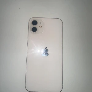 iPhone 12 - Snygg vit iPhone 12 med dubbla kameror och klassisk design. Telefonen ser ut att vara i gott skick med endast mindre spår av användning på kanterna. Perfekt för dig som vill ha en pålitlig och stilren mobil med bra prestanda och kamera.