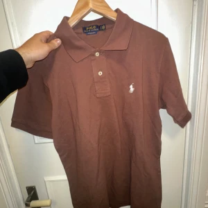 Brun polo från Ralph Lauren - Snygg brun polo från Ralph Lauren med klassisk krage och två knappar framtill. Custom slim fit-modell med det ikoniska broderade logot på bröstet. Skön bomullskvalitet och korta ärmar, perfekt för en chill och stilren look.