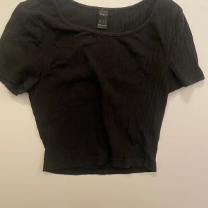 Svart ribbad croppad topp från SHEIN - Svart croppad topp från SHEIN i storlek XS. Toppen har korta ärmar, rund halsringning och är ribbad för en snygg struktur. Perfekt att styla med höga jeans eller kjol för en chill look. Materialet känns mjukt och stretchigt.