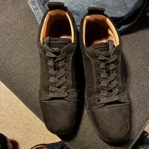 Christian Louboutin svarta sneakers - Säljer ett par svarta sneakers från Christian Louboutin i mocka med klassisk röd sula och broderad logga på hälen. Skorna har snörning, rund tå och läderfoder för extra komfort. Perfekta för dig som vill ha en lyxig och stilren look.