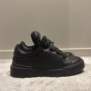 Svarta sneakers Dolce & Gabbana - Snygga svarta sneakers från Dolce & Gabbana med chunky platt sula och breda snören. Skorna är tillverkade i skinn och har en rund tå samt vadderad plös och häl. Märkeslogga på baksidan av sulan. Perfekt för dig som gillar stilrena och lyxiga sneakers. Nypris 12000 mitt pris 1700. Hör av er vid frågor, 