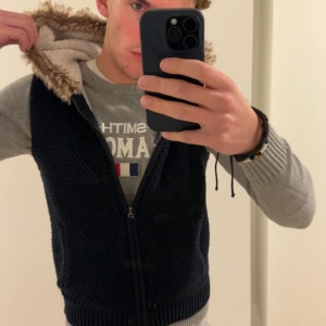 Blå cardigan stickad med pälsluva🐺 - Svart stickad väst med huva, fodrad med mjuk vit fleece och kantad med brun päls. Västen har två fickor fram och stängs med dragkedja. Perfekt för lager-på-lager och chill höststil. Kvinnlig storlek L, manlig storlek S.