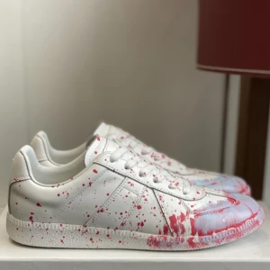 Vit sneaker med röd färgstänk - Unika vita sneakers med coola röda och ljusblå färgstänk över hela skon. Modellen har snörning och platt sula, tillverkad i skinn för en lyxig känsla. Perfekt för dig som vill sticka ut med ett kreativt och konstnärligt uttryck.