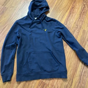 Marinblå hoodie från Lyle & Scott - Snygg marinblå hoodie från Lyle & Scott med gul broderad logga på bröstet. Tröjan har huva med snörning, känguruficka och ribbade muddar. Tillverkad i mjuk bomull som är skön att ha på sig. Perfekt för dig som gillar stilren och enkel design.