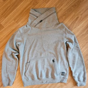 Grå hoodie från Jack & Jones Core - Snygg grå hoodie från Jack & Jones Core med unik hög krage och dragkedja. Tröjan har en stor ficka framtill med extra dragkedja och ribbade muddar. Mjuk insida i fleece och stilren look, perfekt för chill dagar. Priset kan diskuteras!