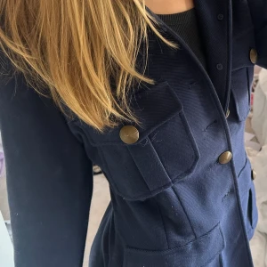 Marinblå kavaj från Polo Ralph Lauren - Snygg marinblå kavaj från Polo Ralph Lauren med markerad midja, fyra stora fickor framtill och mässingfärgade knappar. Jackan är tillverkad i bomull. Helt oanvänd med prislapp kvar nypris: 4895 kr. Äkthets bevis med qr kod i lapp på kavajen/jackan 