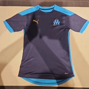 Blå Marseille fotbollströja Puma XS - Säljer en officiell Olympique de Marseille fotbollströja från Puma i storlek XS. Tröjan är mörkblå med ljusblå detaljer på axlar, ärmslut och sidor. Tryckt Marseille-logga och Puma-märke på bröstet. Materialet är lätt och andas, perfekt för träning eller match.