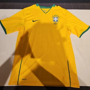 Brasilien fotbollströja Nike M - Gul Brasilien fotbollströja från Nike med gröna detaljer vid krage och ärmslut. Broderat CBF-emblem på bröstet och Nike-logga. Tillverkad i lätt, ventilerande polyester med Dri-FIT-teknologi. V-ringad hals och korta ärmar. Perfekt för dig som älskar brasiliansk fotboll.