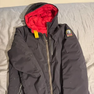Parajumpers vändbar pufferjacka svart/röd - Vändbar pufferjacka från Parajumpers med huva, ena sidan svart och andra sidan röd. Jackan har dragkedja, bröstficka och ikonisk gul detalj vid halsen. Märkespatch på ärmen. Perfekt för dig som vill variera din stil och hålla dig varm. Jackan är äkta 