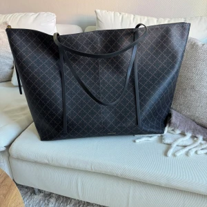 Svart shoppingväska från Malene Birger -  Stor och rymlig shoppingväska från By Malene Birger i svart med diskret rutigt mönster. Väskan har långa svarta axelband, guldfärgad dragkedja och en innerficka. Tillverkad i slitstarkt syntetmaterial och levereras med dustbag. Pris går att diskutera 