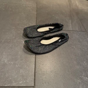 Svarta glittriga ballerinaskor - Säljer ett par svarta ballerinaskor med glittriga detaljer och fyrkantig tå. Skorna har en låg profil och är täckta av små, skimrande stenar som reflekterar i olika färger. Perfekta för dig som vill sticka ut med en snygg och trendig look.