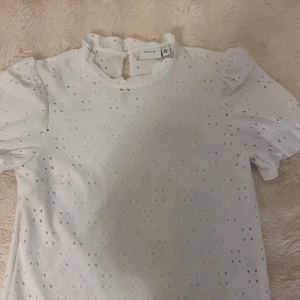 Vit broderad blus från Vila XS - Supersöt vit blus från Vila i storlek XS med broderade hålmönster och puffiga korta ärmar. Blusen har liten volang vid halsen och knapp i nacken för en extra gullig touch. Perfekt för dig som gillar romantisk och feminin stil.