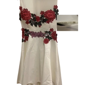 Vit kortklänning med blommig spets - Supersöt vit kortklänning från Meshki med broderade blommor i rött och lila över byst och midja. Klänningen har ärmlös design, markerad midja och lätt utsvängd kjol. Perfekt för dig som vill sticka ut med romantiska detaljer och unik look.