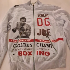 Dolce & Gabbana boxartröja med tryck - Unik ljusgrå ziptröja från Dolce & Gabbana med boxningsinspirerat tryck i svart, rött och brunt. Tröjan har två snedställda dragkedjor framtill, ribbade muddar och coola detaljer. Perfekt för dig som gillar sportiga och statement-plagg.