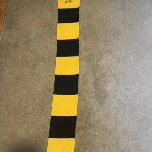 Elfsborg halsduk i svart och gult - Lång halsduk med breda svarta och gula ränder, broderad Elfsborg-logga och svarta fransar i ändarna. Perfekt för att visa ditt stöd på matchen och hålla dig varm. Tjockt stickat material som känns mjukt och skönt runt halsen.