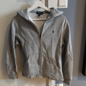 Grå hoodie från Polo Ralph Lauren - Snygg grå hoodie från Polo Ralph Lauren med dragkedja och huva. Klassisk design med broderad logga på bröstet och två fickor framtill.