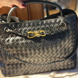 Bottega Veneta inspired Svart flätad handväska med gulddetalj - Snygg svart handväska i flätat skinn med guldfärgad metallspänne framtill. Väskan har ett stilrent och elegant utseende med praktiskt handtag och flätade detaljer. Perfekt för dig som gillar klassisk design med en twist.