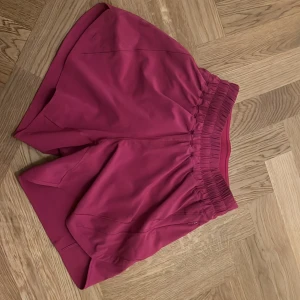 Rosa Hotty hot träningsshorts från Lululemon - Säljer ett par super snygga, rosa träningsshorts från Lululemon. De har hög midja med bred resår och snörning, samt diskreta fickor med dragkedja på sidorna. Perfekta för gymmet eller löprundan, tillverkade i ett mjukt och stretchigt material.