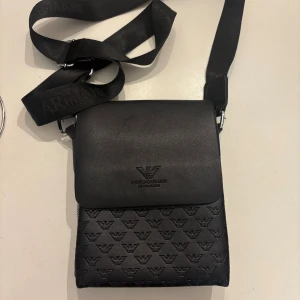 Svart axelväska från Giorgio Armani - Snygg svart axelväska från Giorgio Armani med präglad logotyp på framsidan och justerbar axelrem. Väskan har flera fack, dragkedja bak och invändigt samt diskret mönster med Armani-loggor. Tillverkad i skinnliknande material.