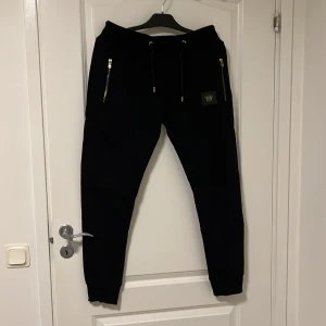 Svarta mjukisbyxor från MR Originals - Svarta mjukisbyxor från MR Originals med gulddetaljer på dragkedjor och snörning i midjan. Byxorna har smal passform och ribbade muddar vid bensluten. Märkeslogga med krona på vänster ben. Perfekta för en chill och stilren look.