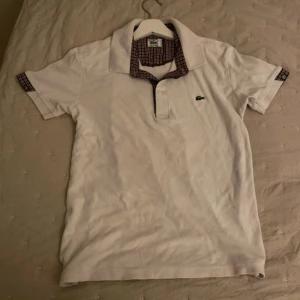 Vit pikétröja Lacoste med rutiga detaljer - Vit pikétröja från Lacoste med klassisk krage och rutigt tyg på insidan av kragen och ärmsluten. Tröjan har korta ärmar och den ikoniska gröna krokodilloggan broderad på bröstet. Perfekt för en clean och stilren look.