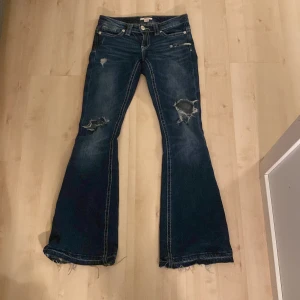 Bootcut jeans från Gina Tricot XS - Mörkblå bootcut jeans från Gina Tricot i storlek XS. Jeansen har slitna detaljer och hål på knäna, fransiga benslut och snygga broderade bakfickor. Klassisk femficksmodell med låg midja och dragkedja. Perfekta för en cool och avslappnad look.