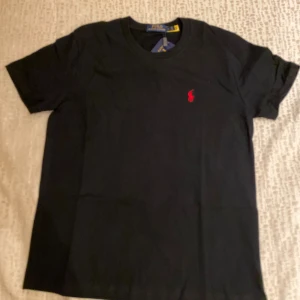 Svart t-shirt från Polo Ralph Lauren - Svart t-shirt från Polo Ralph Lauren med klassisk röd broderad logga på bröstet. T-shirten är i bomull, har rund hals och korta ärmar. Enkel och stilren modell som passar perfekt till jeans eller shorts.