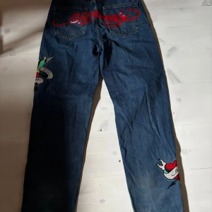 Ed Hardy jeans med broderier, W26 - Mörkblå jeans från Ed Hardy med färgglada broderier och tryck, bland annat ett hjärta och fågel på benen samt röd Ed Hardy-logga bak. Klassisk femficksmodell i bomullsjeans med raka ben och råa kanter nertill för en edgy vibe.
