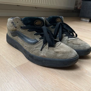 Olivgröna Zahba Beatrice Domond X Vans mid - Unika Vans Beatrice sneakers i grön mocka med svarta detaljer och snörning. Skorna har en klassisk Vans-sida i svart skinn och svart platt sula. Insidan och plösen är svarta, och namnet Beatrice är broderat i guld på hälen. Perfekt för dig som gillar skate och streetstyle. Innersula med vr3 cush för bra dämpning