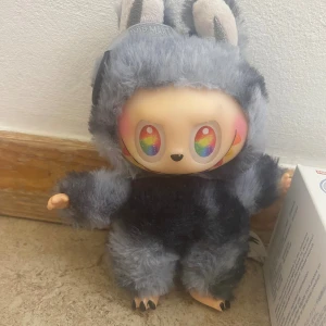 Pop Mart Biginto Energy Vinyl Plush Pendant Blind Box - Söt Pop Mart Biginto Energy vinyl plush figur med regnbågsfärgade ögon och mjuk grå pälsdräkt. Kommer med originalförpackning. Perfekt skick, inga synliga defekter eller slitage. Samlarobjekt och dekorativ accessoar.