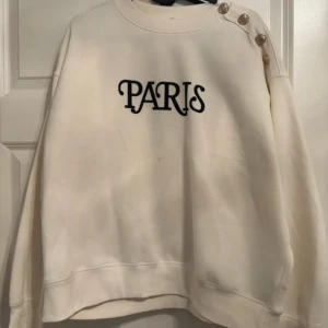 Vit sweatshirt med PARIS-tryck och knappar - Säljer en oversized vit sweatshirt med svart PARIS-tryck på bröstet. Tröjan har snygga guldknappar längs ena axeln som cool detalj. Mjuk och skön, perfekt till jeans eller kjol. Rund halsringning och långa ärmar.