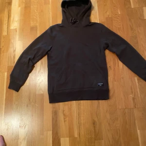 Mörkbrun hoodie från Björn Borg - Snygg mörkbrun hoodie från Björn Borg med klassisk känguruficka och huva med snörning. Tröjan har en stilren look och en liten svart etikett med logga på fickan. Perfekt för chill dagar och enkel att matcha med jeans eller joggers.