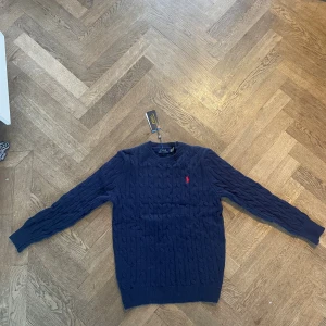 Marinblå stickad tröja från Polo Ralph Lauren - Snygg marinblå stickad tröja från Polo Ralph Lauren med klassiskt kabelmönster och röd broderad logga på bröstet. Rund hals och långa ärmar, perfekt för dig som gillar stilrena och tidlösa plagg.