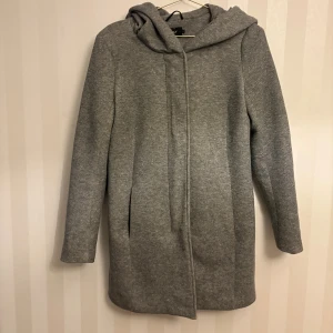 Grå kappa med huva från ONLY - Snygg grå kappa från ONLY med huva och dold dragkedja samt knappar framtill. Jackan har lång ärm och är tillverkad i ett mjukt material som känns både stilrent och bekvämt. Perfekt för kyligare dagar när du vill hålla stilen enkel och clean.