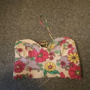 Blommig korsett med snörning - Superfin korsettopp med färgglatt blommönster i rosa, gul, grön och lila. Toppen har smala axelband och snygg snörning både fram och bak för en justerbar passform. Perfekt för dig som vill sticka ut med en retrovibe och coola detaljer.