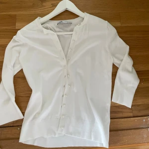 Vit topp med pärlknappar från Zara - Säljer en stilren vit blus från Zara med trekvartsärm och v-ringning. Blusen har dekorativa pärlknappar längs hela framsidan och är tillverkad i ett mjukt material som känns skönt mot huden. Perfekt för dig som gillar en clean och elegant look.