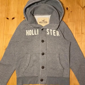 Grå hoodie med knappar från Hollister - Snygg grå hoodie från Hollister med stora vita bokstäver över bröstet. Tröjan har huva, knäppning med knappar framtill och två fickor. Perfekt för en chill och avslappnad stil.