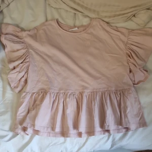 Ljusrosa volangblus från Kappahl - Supersöt ljusrosa blus från Kappahl med volanger både på ärmarna och nertill. Blusen har rund halsringning och är tillverkad i mjuk bomull. Perfekt för dig som gillar en romantisk och trendig look med extra volym på ärmarna. Ny och alldig använd därefter priset ❤️