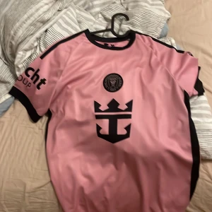 Rosa Inter Miami CF fotbollströja M - Säljer en rosa fotbollströja från Inter Miami CF med svarta detaljer och klubbmärke på bröstet. Tröjan har korta ärmar, svart krage och är tillverkad i ett lätt, sportigt material. Stor logga på framsidan och tryck på ärmen.