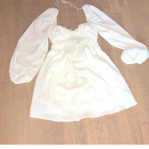 Vit kort klänning från Bershka - Superfin vit klänning från Bershka med puffiga långärmar och söt rynkad detalj vid bysten. Klänningen har en kort modell och är perfekt för dig som gillar romantisk och trendig stil. Materialet känns lätt och luftigt, och den har en snygg fyrkantig ringning.