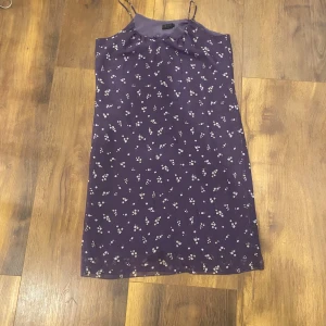 Lila blommig sommarklänning - Söt lila klänning med smala axelband och små vita blommor över hela tyget. Rak passform och lätt, luftig känsla – perfekt för varma dagar. Klänningen är knälång och har en enkel, stilren design som känns både trendig och avslappnad.